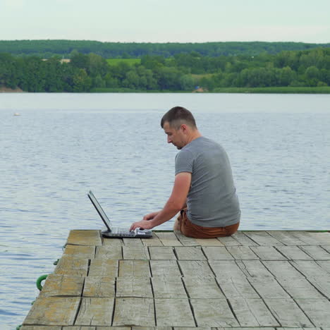 handsome man sitting at the edge of the bridge finishes typing on a laptop and enjoys the natural background. work with pleasure outdoors near the river