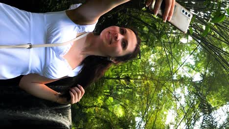 vertical ascending shot of a young attractive influencer taking a selfie in front of a waterfall in bali indonesia for social media in the jungle