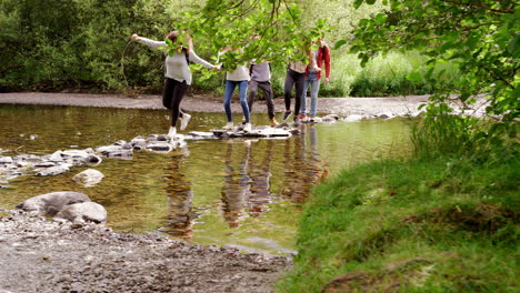 Cinco-Amigos-Adultos-Jóvenes-Se-Toman-De-La-Mano-Y-Se-Ayudan-Mutuamente-A-Cruzar-Un-Arroyo-Balanceándose-Sobre-Piedras-Durante-Una-Caminata