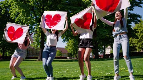 mujeres jóvenes amigas que se unen, saltando juntas en el parque verde, sosteniendo carteles blancos de gran tamaño decorados con corazones rojos vibrantes, irradiando felicidad compartida y espíritu de equipo