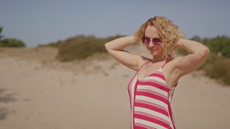 a blonde woman stands with her arms behind her head and waves her hair in slow motion, basking in the sun atop bright white sand dunes.