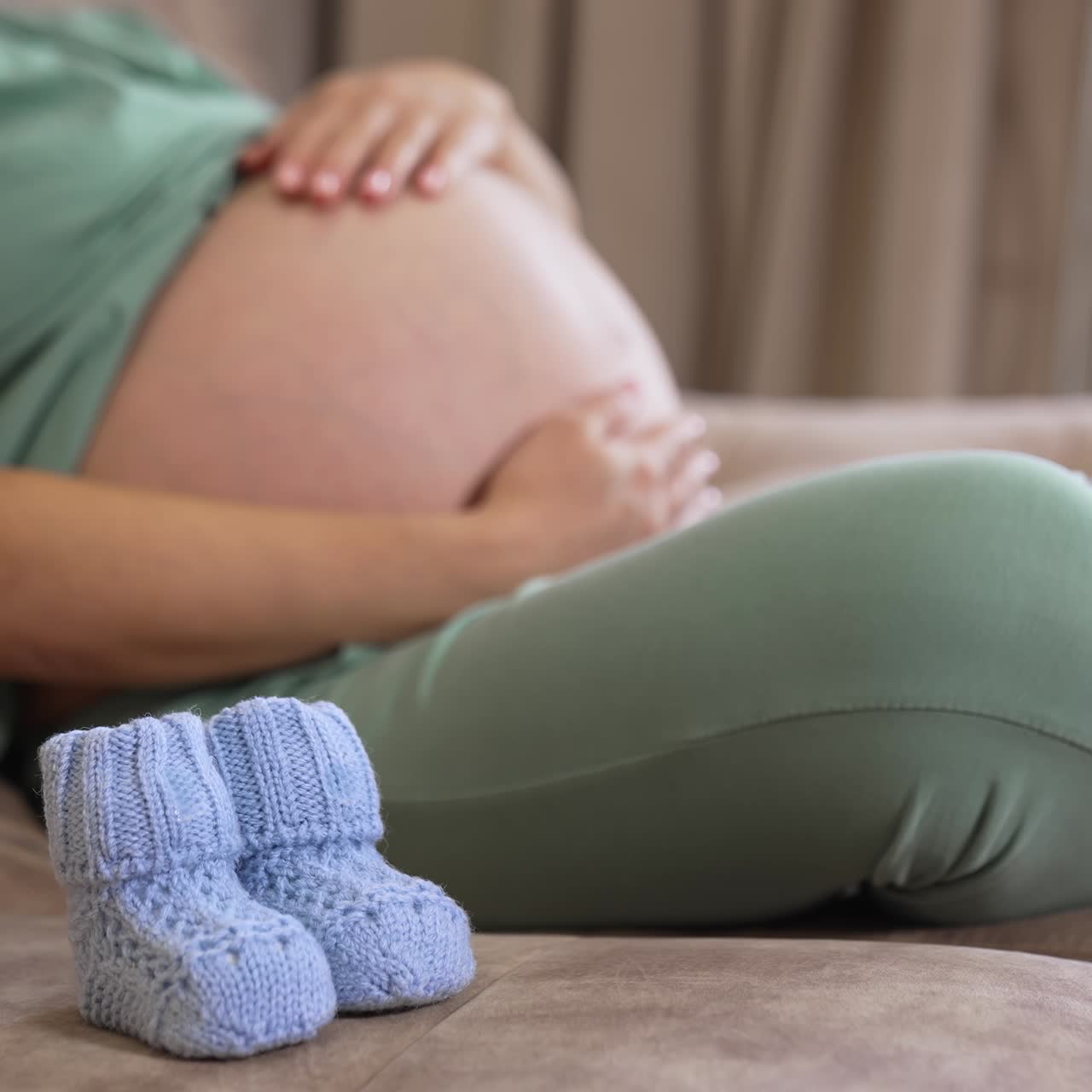Pregnant woman with big bare belly sitting on the couch. Lady caressing her pregnant belly tenderly and then putting baby socks on it