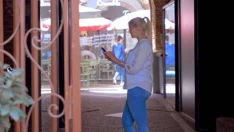 behind a red barrier, a blonde caucasian woman walks up, stops to take out her smartphone and take a picture of the window on the left