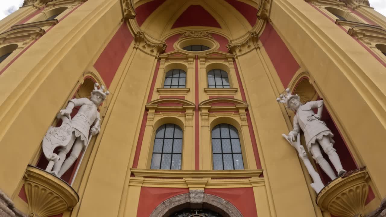Camera tilts down the ornate red and yellow baroque front of Stift Wilten in Innsbruck, Austria, highlighting two white warrior statues set in niches above the arched church entrance