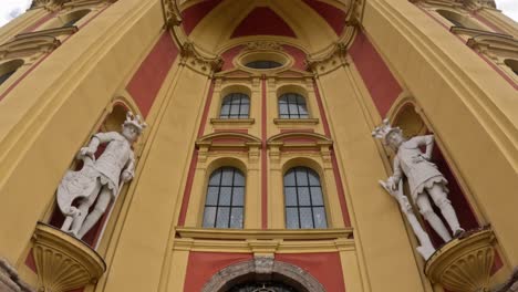camera tilts down the ornate red and yellow baroque front of stift wilten in innsbruck, austria, highlighting two white warrior statues set in niches above the arched church entrance