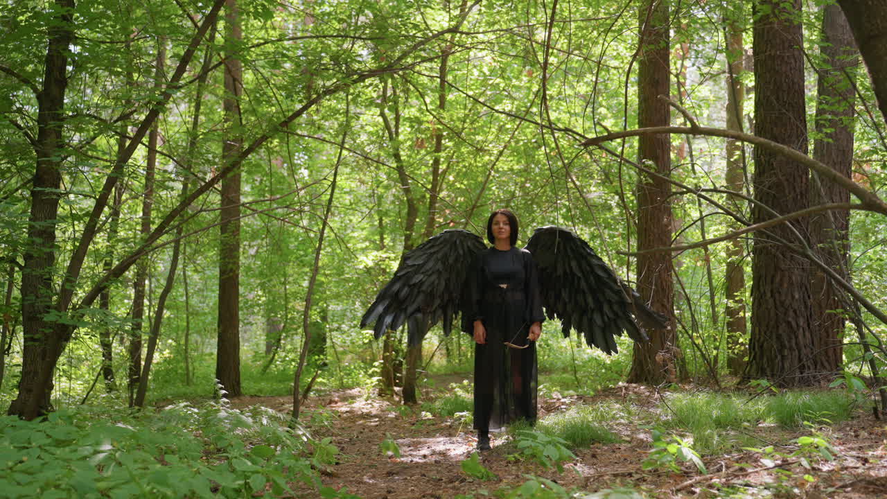 Black fairy goddess with large dark wings stands gracefully in lush green forest, holding bow as sunlight filters through leaves and tiny tree particles drift around