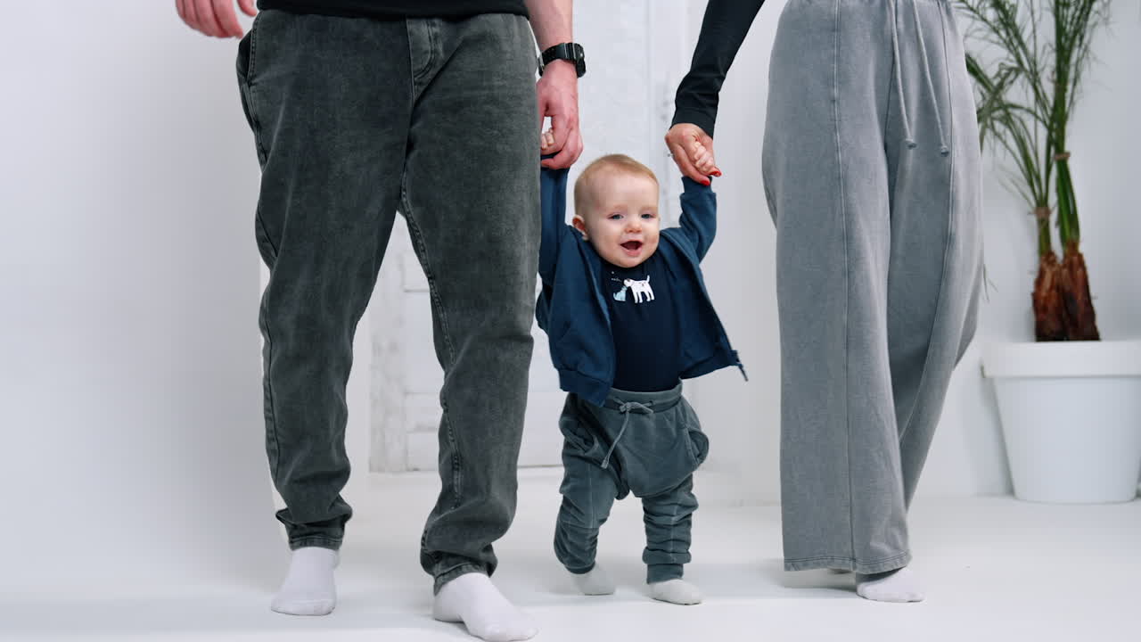 Lovely infant wearing jeans making first awkward steps. Unrecognized parents support their son.