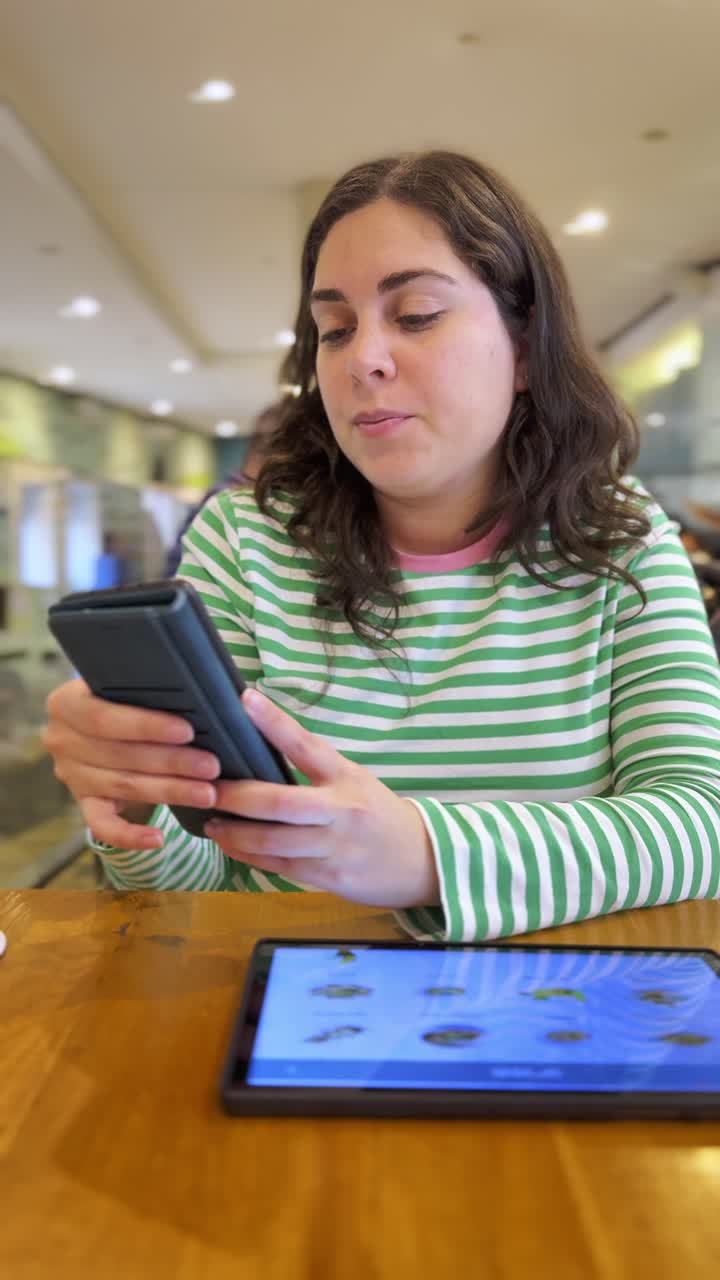A woman is sitting at a table with a tablet and a cell phone