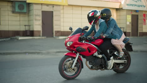 two women ride a red power bike through an urban street, with the rider's button-down shirt open, they pass by a building, with sunlight reflecting off them