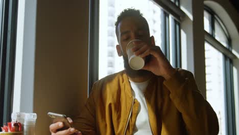 Front-view-of-young-mixed-race-man-having-coffee-while-using-mobile-phone-in-cafeteria-4k