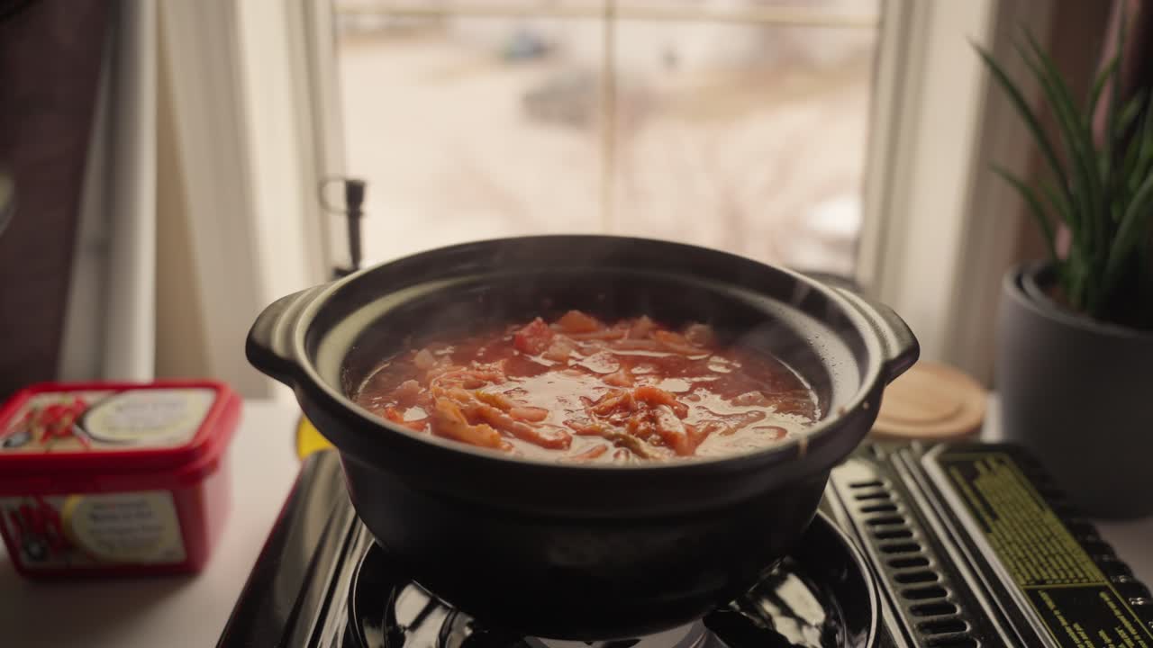 Un cucchiaio di legno mescola una zuppa che sobbolle all'interno di una pentola di ceramica su un fornello portatile. Il vapore sale mentre gli ingredienti cuociono in un ambiente caldo di una cucina domestica.