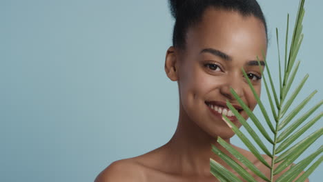 retrato de una mujer afroamericana atractiva tocando la cara con una hoja acariciando una piel hermosa y saludable disfrutando de la belleza natural juguetona sobre un fondo azul