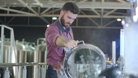 young male brewer wearing a leather apron supervise the process of beer fermentation at a modern brewery factory