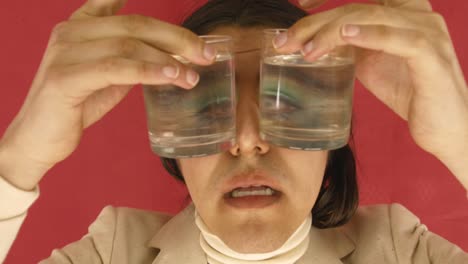 Man-In-Red-Background-Holding-Two-Glasses-Of-Water-On-His-Eyes---close-up