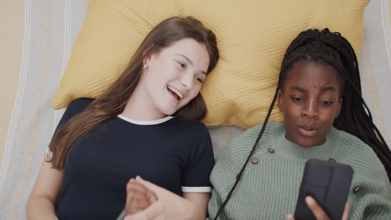 felices amigas adolescentes diversas acostadas en la cama usando teléfonos inteligentes y riendo