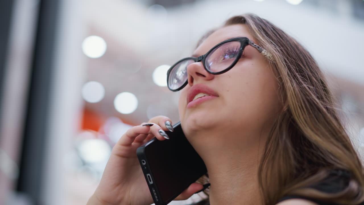 primer plano de una mujer hermosa hablando por teléfono con camiseta negra y gafas, mirando a su alrededor, con fondo borroso con efecto de luz bokeh y estructuras modernas