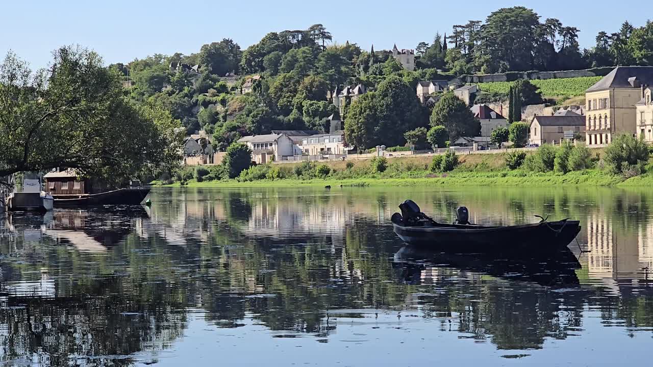 A static shot shows a rowboat on the river with gentle water movement. Other boats appear in the distance, and Chinon town with trees and reflections is visible on the opposite bank