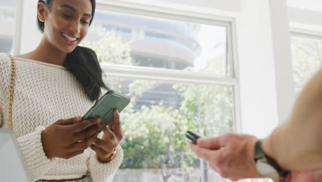 Feliz-Mujer-Birracial-Pagando-A-Un-Barista-Masculino-Caucásico-Usando-Un-Teléfono-Inteligente-En-La-Cafetería