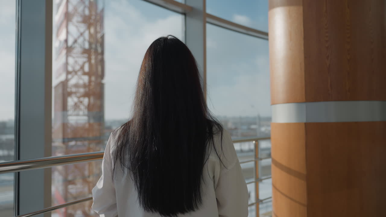 Rear view of woman in white shirt with long dark hair walking inside modern building with large glass windows overlooking blurry urban landscape under natural daylight