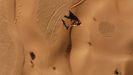 the drone is flying topdown following a man with a camel and a person on top in the dessert with their shadow on the ground in tunisia aerial footage 4k