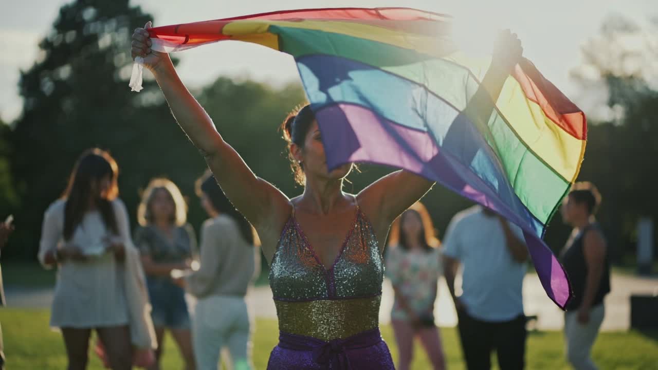 Lifting woman raising rainbow flag in park, sun revealing sparkly top, celebrating pride