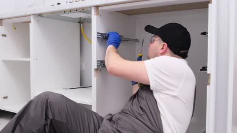 focused handyman in overalls sitting on floor while assembling kitchen