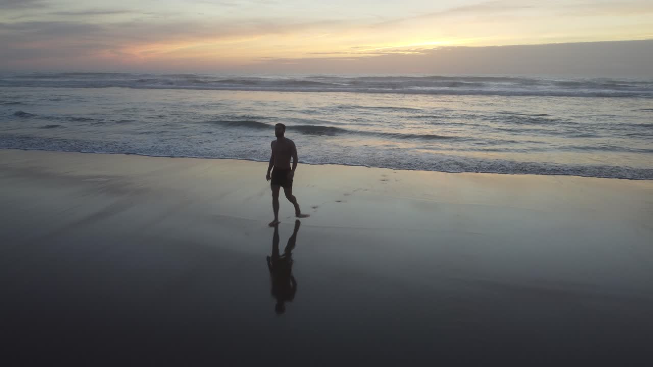 Dynamic Drone shot of a fit man walking out of the water on a cold day, on a beach in Costa da Caparica, Portugal, during the sunset.