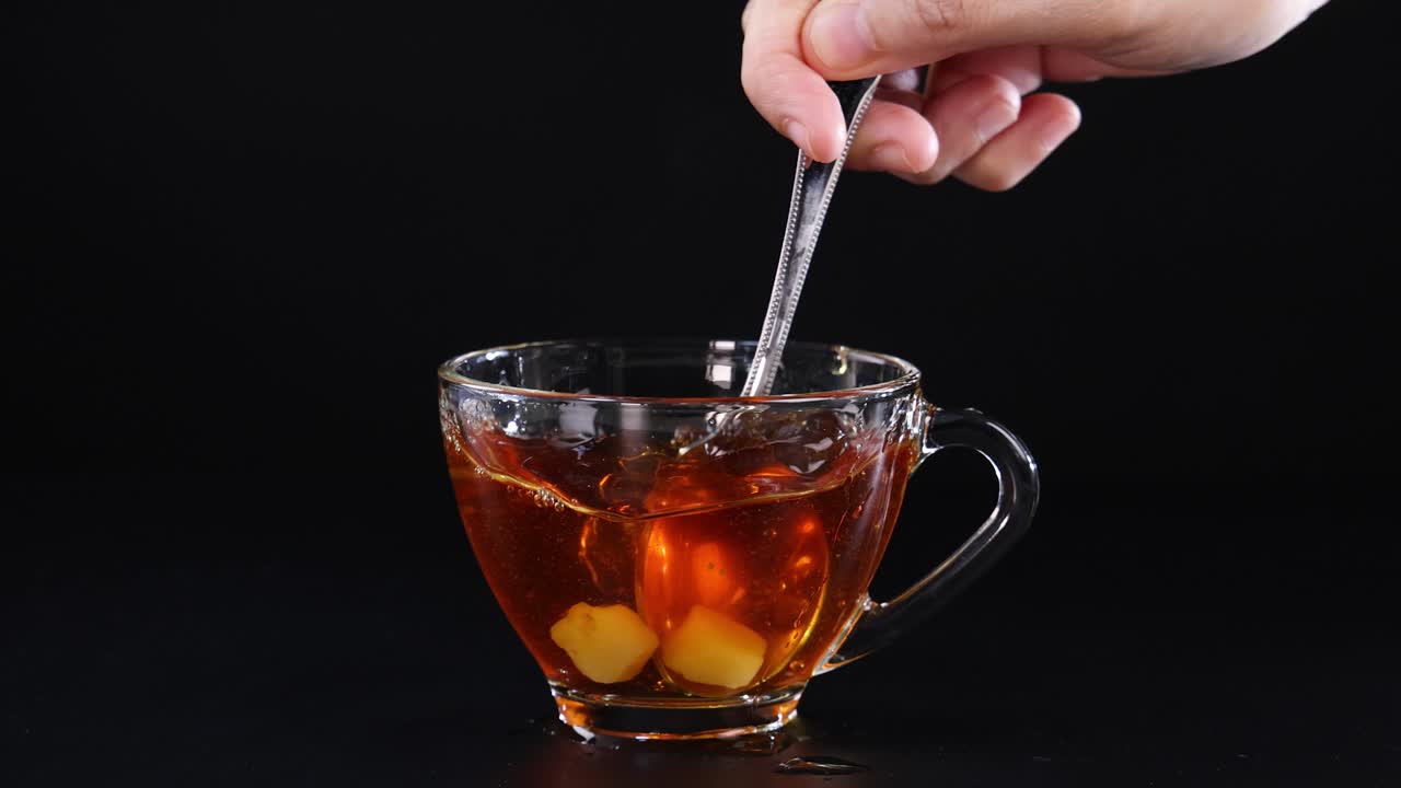 A hand stirs a sugar cube in a glass cup of tea using a spoon against a black background