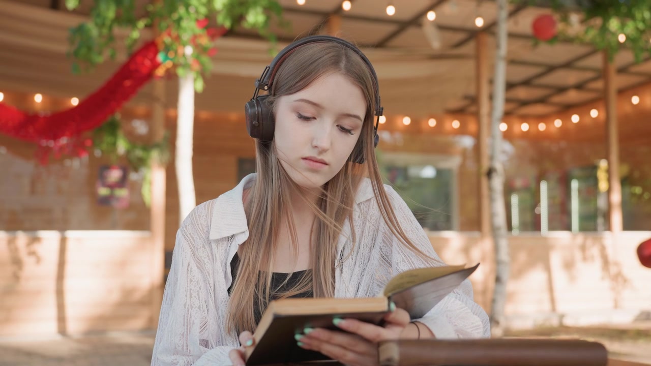 Nærbilde av en ung kvinne med hodetelefoner som leser en bok ved et trebord i lyse utendørs omgivelser med lyslenker og grøntområder. Kvinnen holder en åpen bok omgitt av flere bøker.