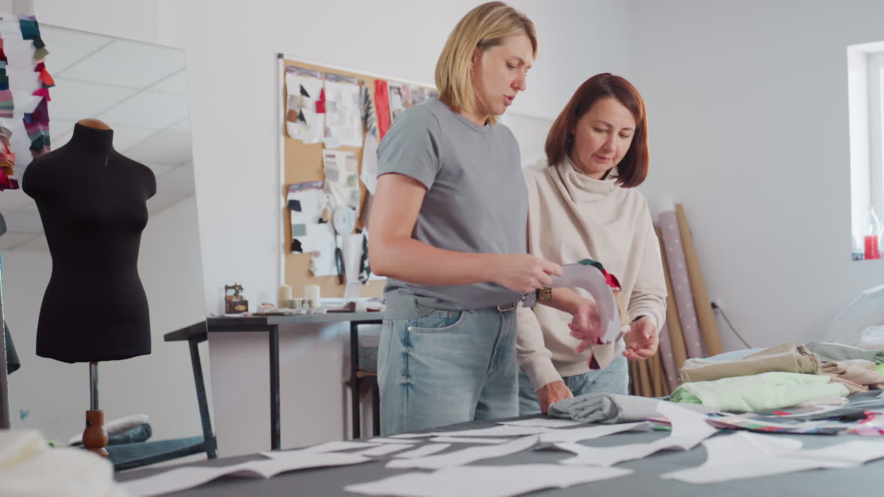 Two women in casual jeans attire walk to workspace and attentively observe colorful fabric samples together on cluttered worktable, surrounded by sewing materials, and inspiration boards