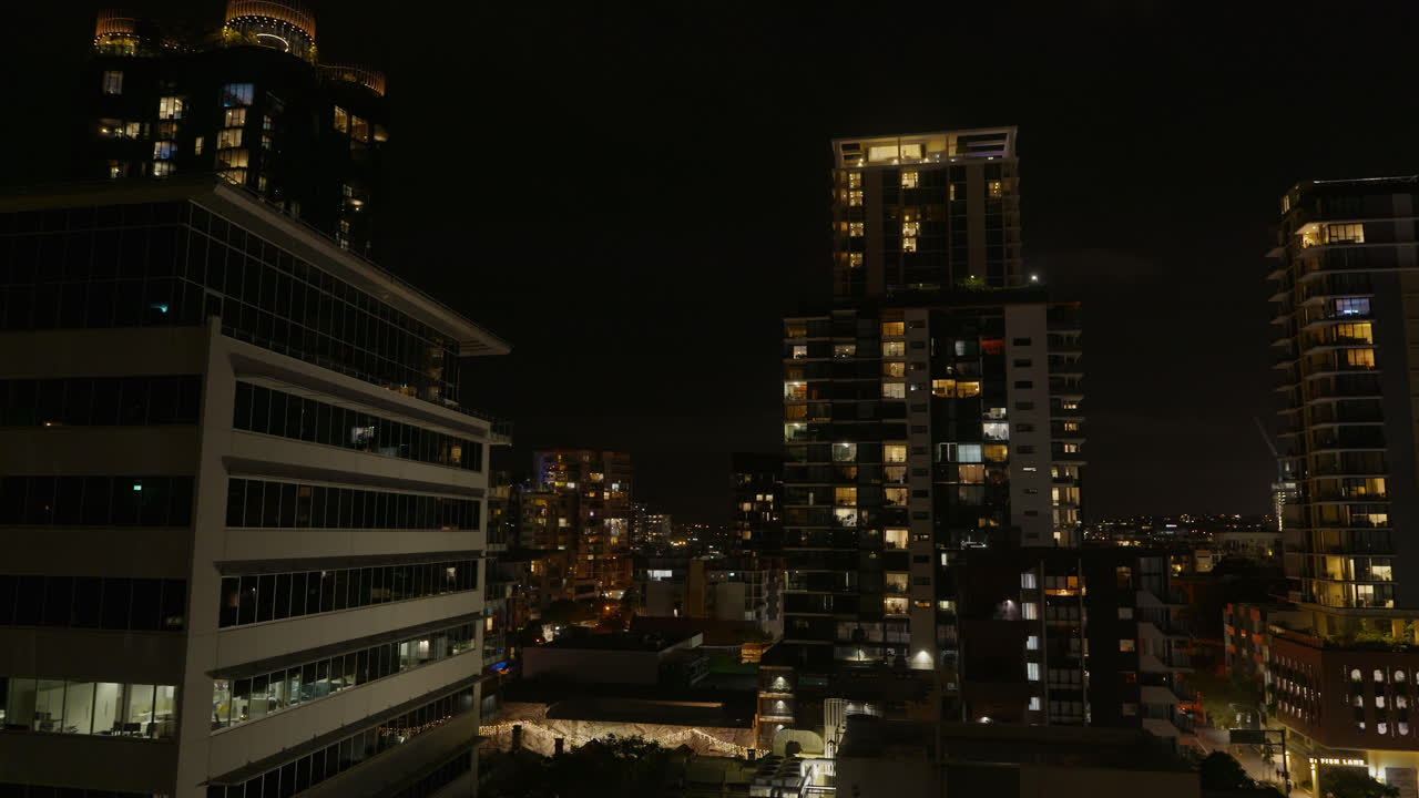 Panorâmica para a direita, edifícios da cidade de Brisbane e arranha-céus à noite com luzes de quartos iluminados
