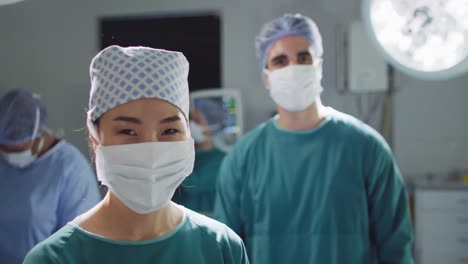 video portrait of asian female surgeon in face mask smiling in operating theatre, with copy space