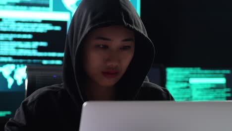 close up of asian female hacker in a hood looking at camera while works on a laptop with maps and data on display screens of multiple monitors in a dark office room