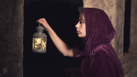 young mysterious woman wearing a cloak and hood holding a lantern looking for something in the dark at night