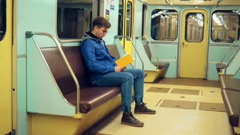 Young man with glasses is reading a book in an empty subway car. Brutal guy rides and reads a book.