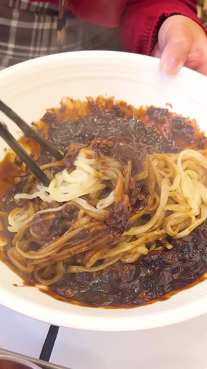 A person mixes Jajangmyeon noodles in a restaurant setting, showcasing the rich, dark sauce and vibrant dining atmosphere
