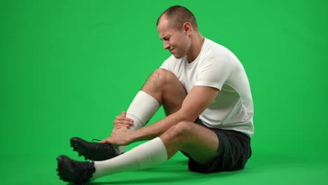 troubled caucasian sportsman sitting on green screen holding injured leg. side view wide shot of sad young handsome man with painful sport trauma at chromakey background.