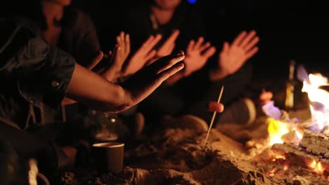 Diverse-group-of-young-men-and-women-sitting-by-the-bonfire-in-the-evening,-warming-up-their-hands-and-talking