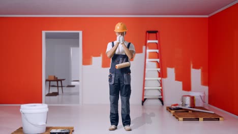 full body of asian man painter wearing safety helmet praying for something while standing with painting wall in room