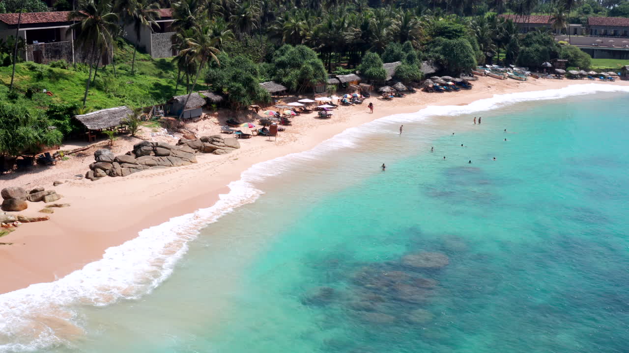 Drone flies backward showing people swimming and enjoying the clear waters near Silent Beach, Sri Lanka.