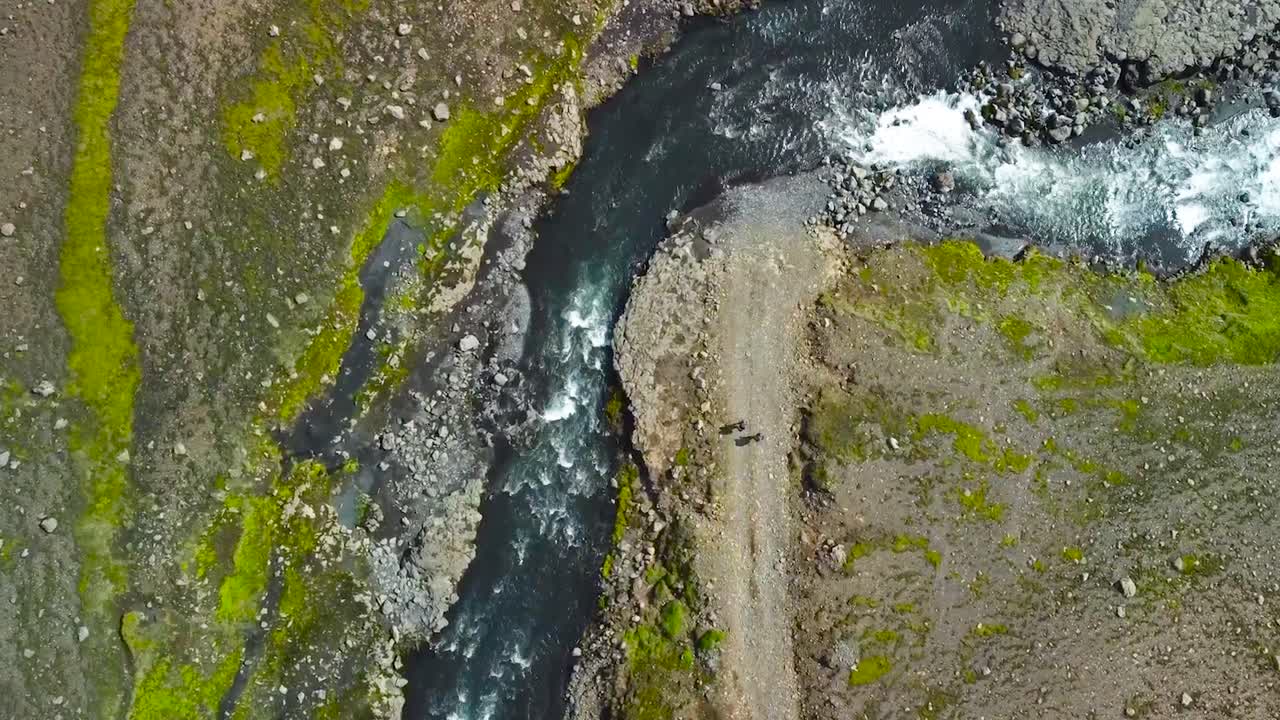 Top down aerial drone footage of two hikers walking on a road or a rural mossy green and brown volcanic Iceland landscape near a fast flowing river during a sunny day. The river turns to the right.