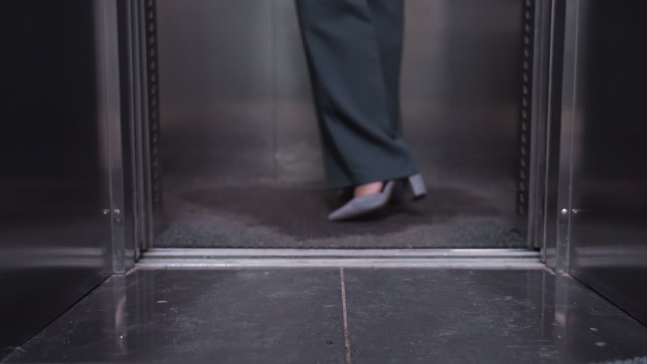 Person stepping out of elevator wearing business attire, focus on legs and shoes, close-up shot showing steps in professional setting, office, movement in modern building interior