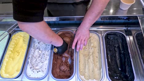 overhead view of a gloved worker scooping chocolate ice cream from a display case with multiple flavors under bright, even lighting in a modern shop