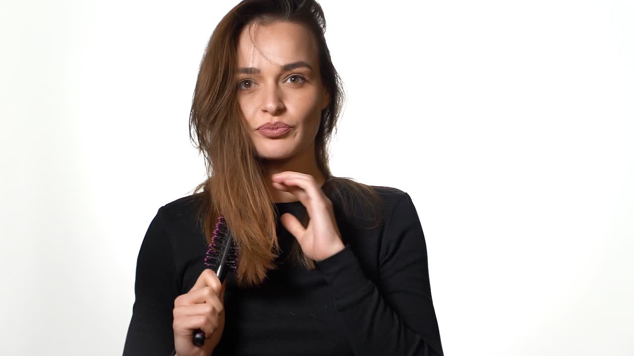 A pensive woman with long hair is looking straight and parallelly combing her hair on a white background. Close-up.