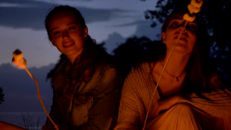 two young caucasian girls sit in the evening by the fire in nature, cooking marshmelow, smiling, eating