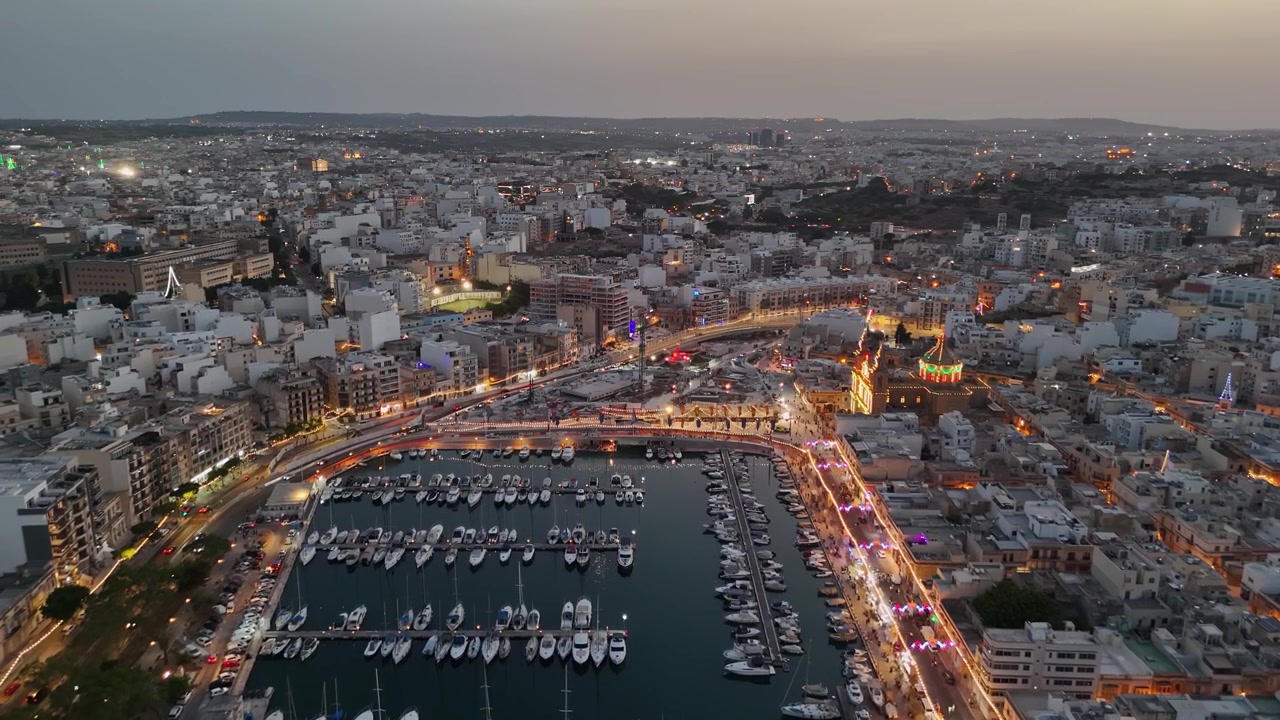 Pemandangan udara tepi laut Msida, Malta, dan Knisja Parrokkjali tal-Imsida saat festa desa di senja hari, dengan kerumunan, perahu, dan pemandangan kota Mediterania yang diterangi.