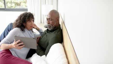 Happy-mature-diverse-couple-sitting-on-bed-using-tablet-and-talking-at-home,-slow-motion