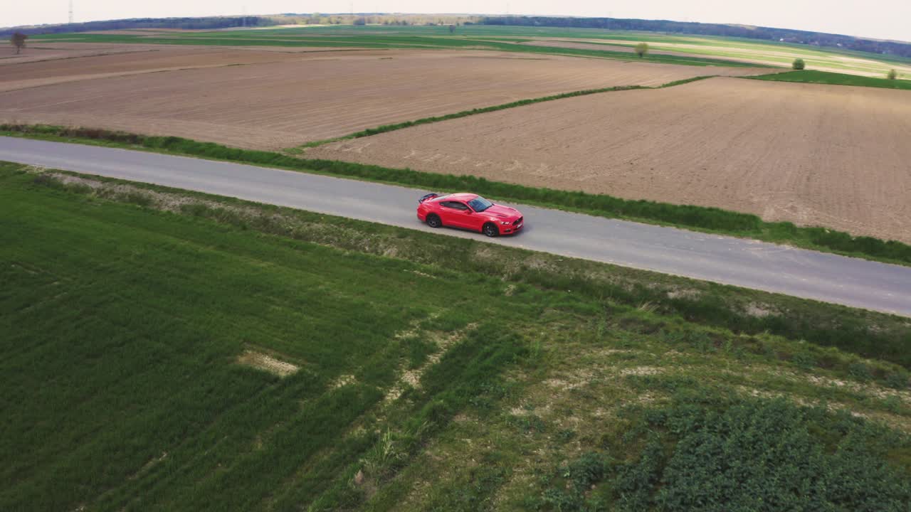 toma cinematográfica de un drone de un ford mustang gt naranja conduciendo por una carretera recta rodeada de campos verdes.