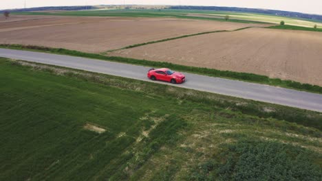 Toma-Cinematográfica-De-Un-Ford-Mustang-Gt-Naranja-Conduciendo-Por-Una-Carretera-Recta-Rodeada-De-Campos-Verdes