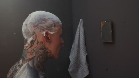 tattooed inmate shaving his head with disposable razor in prison cell